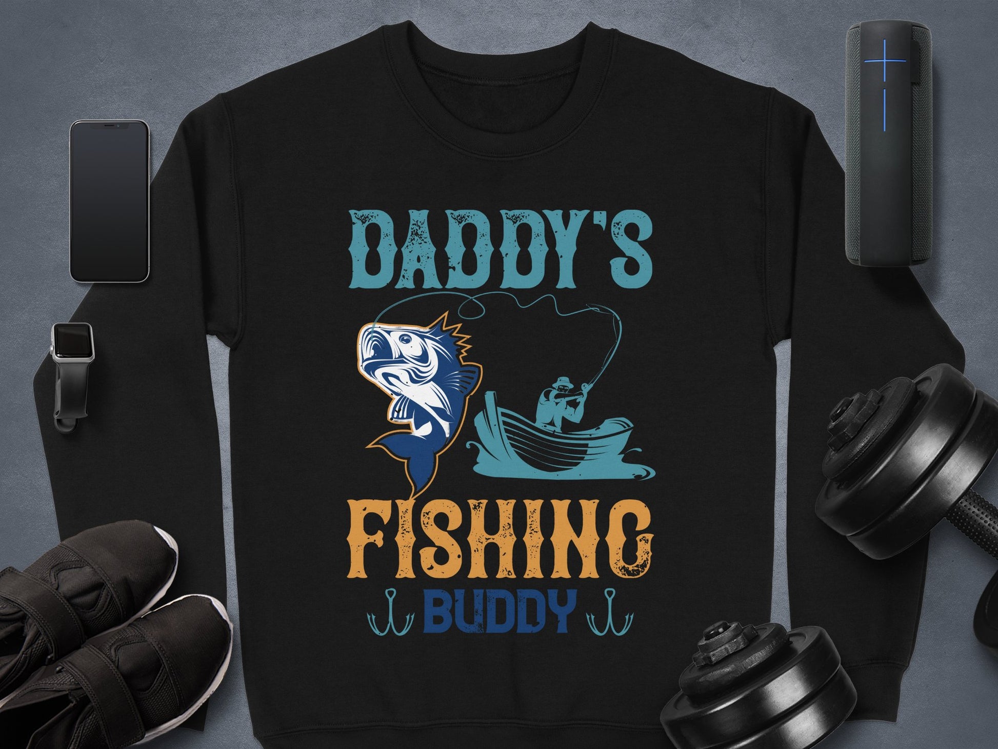 Garment Graphics black sweatshirt features Daddys Fishing Buddy text with a fish and angler illustration. Ideal for outdoor enthusiasts, it is displayed on a gray surface next to a smartphone, smartwatch, shoes, dumbbells, and portable speaker.