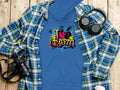 A Garment Graphics blue hoodie with I Love Graffiti and vivid designs layers under a blue-green plaid shirt. Nearby, black headphones, a camera, and a white cup on wood capture the essence of an urban street art-inspired casual style.