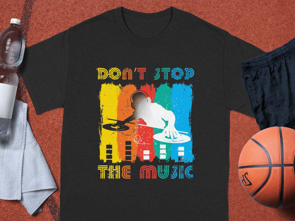 The Garment Graphics black T-shirt showcases a vivid DJ graphic with Don’t Stop the Music in bold, rainbow letters. Ideal for music enthusiasts, it sits on a sports court with a basketball, shorts, towel, and water bottle—perfect for energetic party-goers.