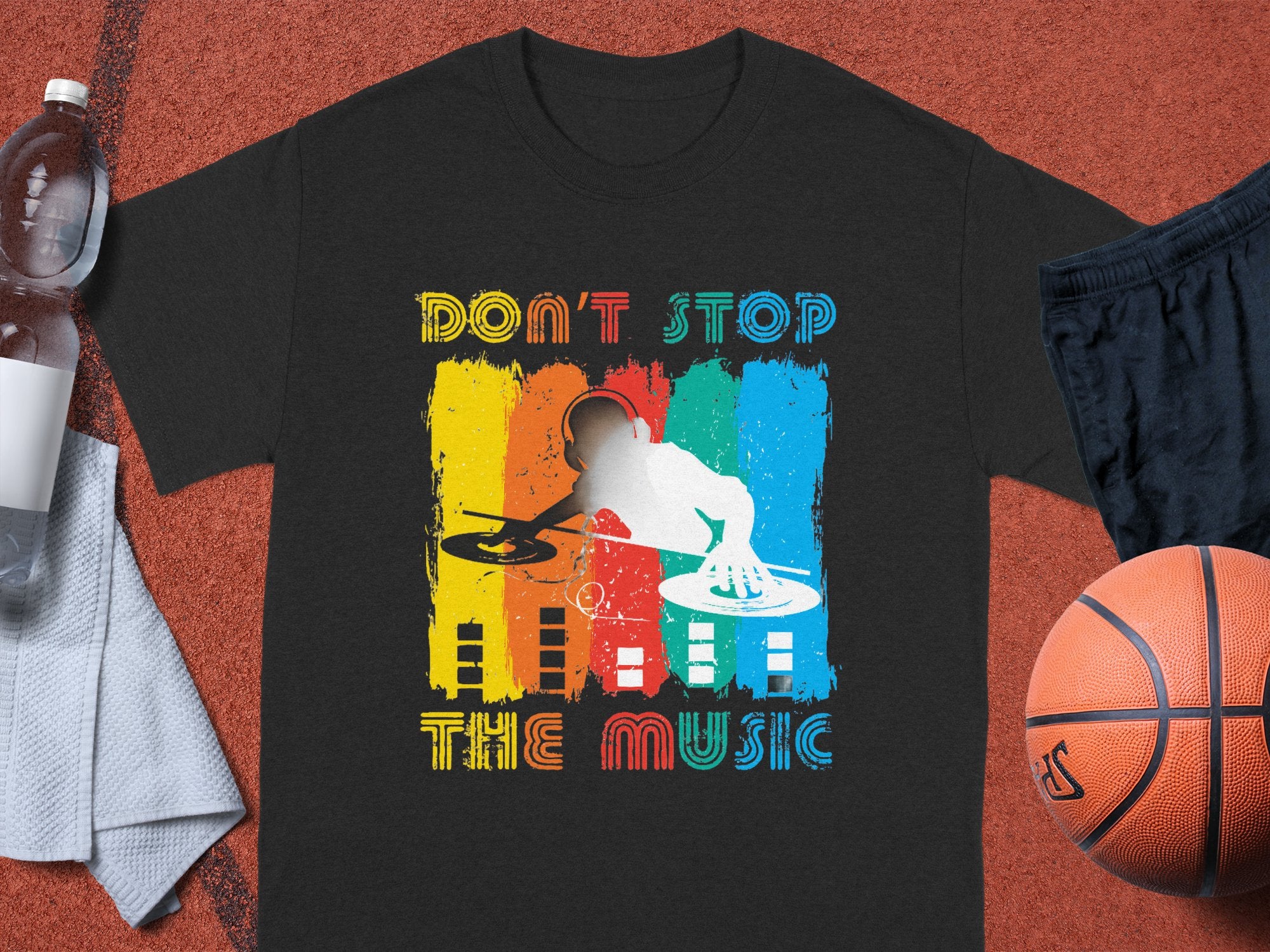 The Garment Graphics black T-shirt showcases a vivid DJ graphic with Don’t Stop the Music in bold, rainbow letters. Ideal for music enthusiasts, it sits on a sports court with a basketball, shorts, towel, and water bottle—perfect for energetic party-goers.