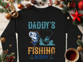 A black Garment Graphics sweatshirt for fishing enthusiasts features Daddys Fishing Buddy with a boat illustration, surrounded by Christmas decor and a coffee cup on a wooden background, ideal for cozy holiday moments.