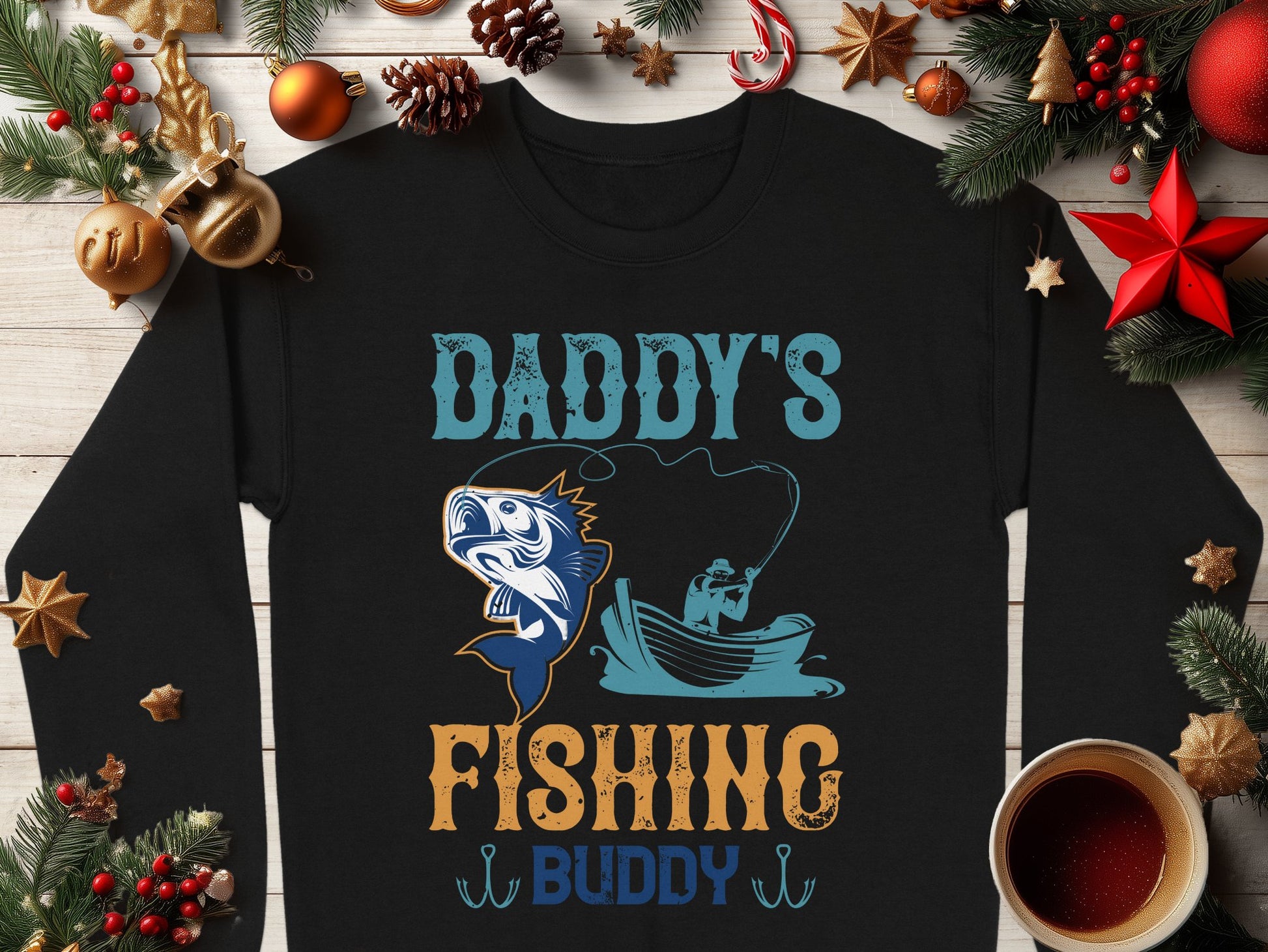 A black Garment Graphics sweatshirt for fishing enthusiasts features Daddys Fishing Buddy with a boat illustration, surrounded by Christmas decor and a coffee cup on a wooden background, ideal for cozy holiday moments.