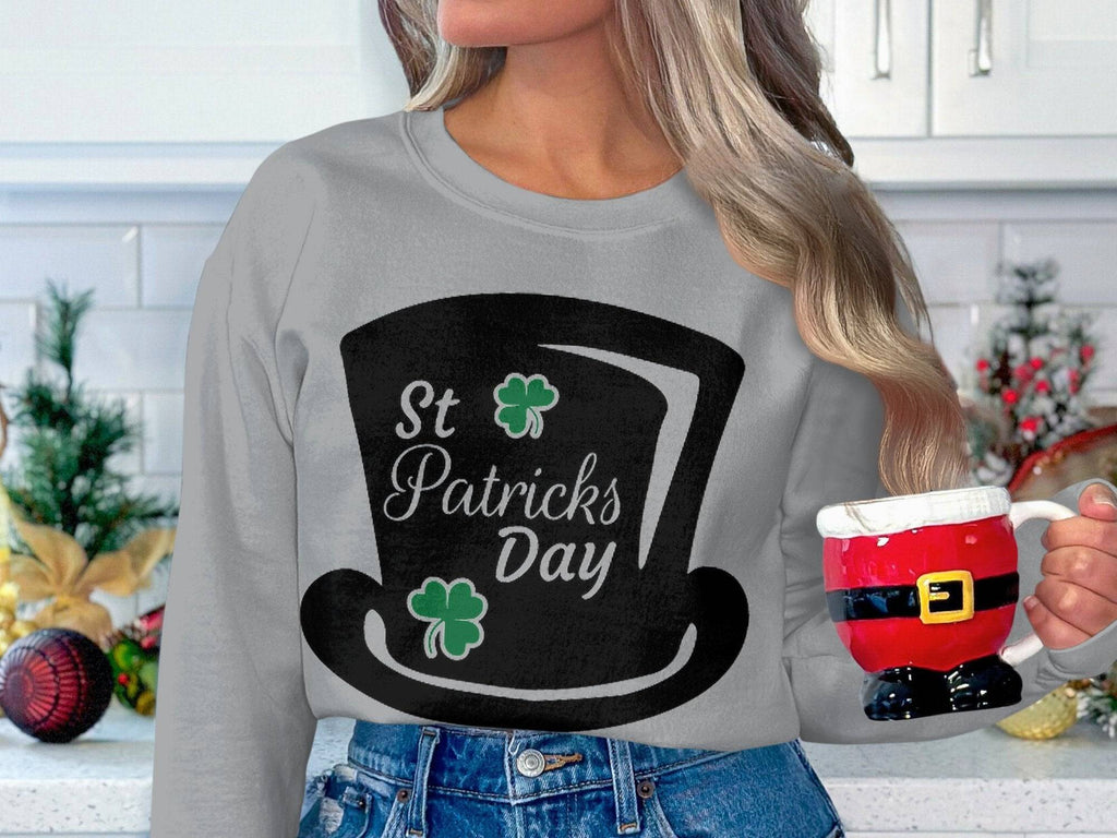 A person with long hair holds a Santa-themed mug, embodying the festive spirit. They wear a stylish gray Garment Graphics sweatshirt featuring a large black top hat with two clovers and St. Patricks Day text against a backdrop of kitchen cabinets and Christmas decorations.