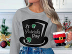 A person with long hair holds a Santa-themed mug, embodying the festive spirit. They wear a stylish gray Garment Graphics sweatshirt featuring a large black top hat with two clovers and St. Patricks Day text against a backdrop of kitchen cabinets and Christmas decorations.