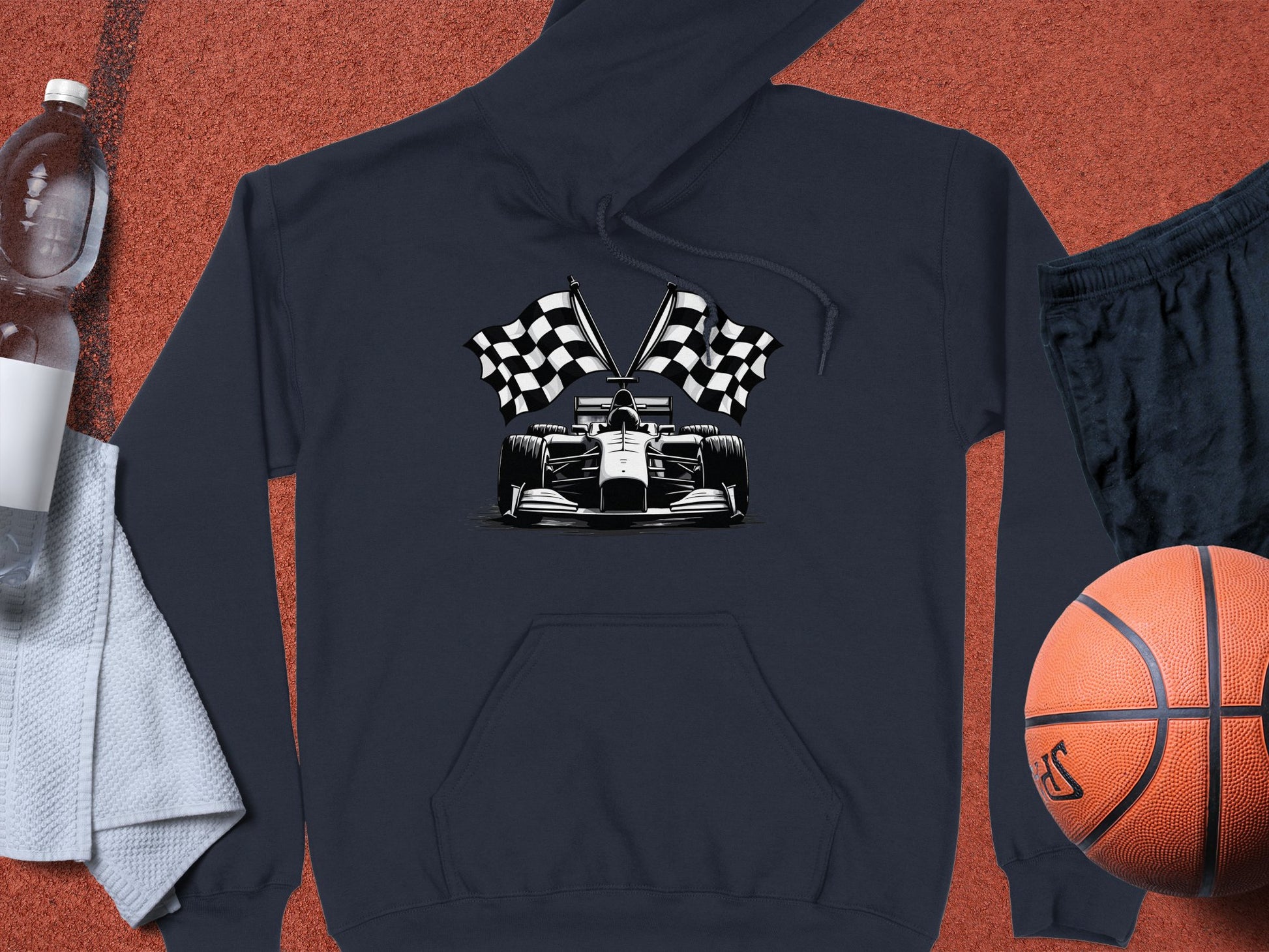 A Garment Graphics racing-themed dark hoodie with a race car and checkered flags lies on a textured surface, accompanied by a basketball, dark shorts, a clear water bottle, and a white towel.