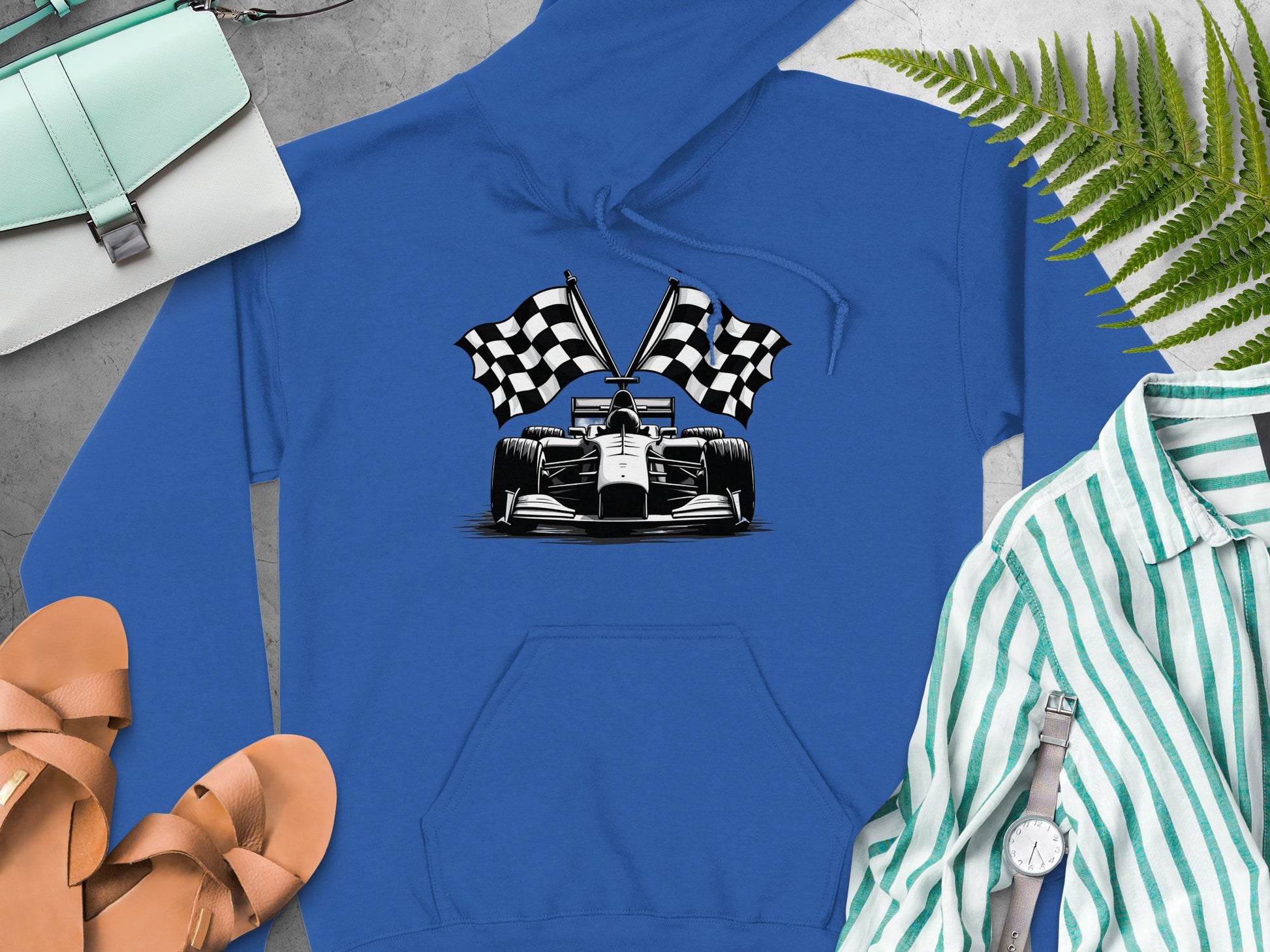 A Garment Graphics blue hoodie with a race car and checkered flags sits on a grey surface, accompanied by a light blue purse, brown sandals, green ferns, and a green and white striped shirt with a watch and bracelet.