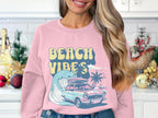 Wearing Garment Graphics pink retro sweatshirt, featuring a classic car, surfboard, wave, palm trees, and Beach Vibes, a person stands in a kitchen adorned with holiday ornaments and greenery.