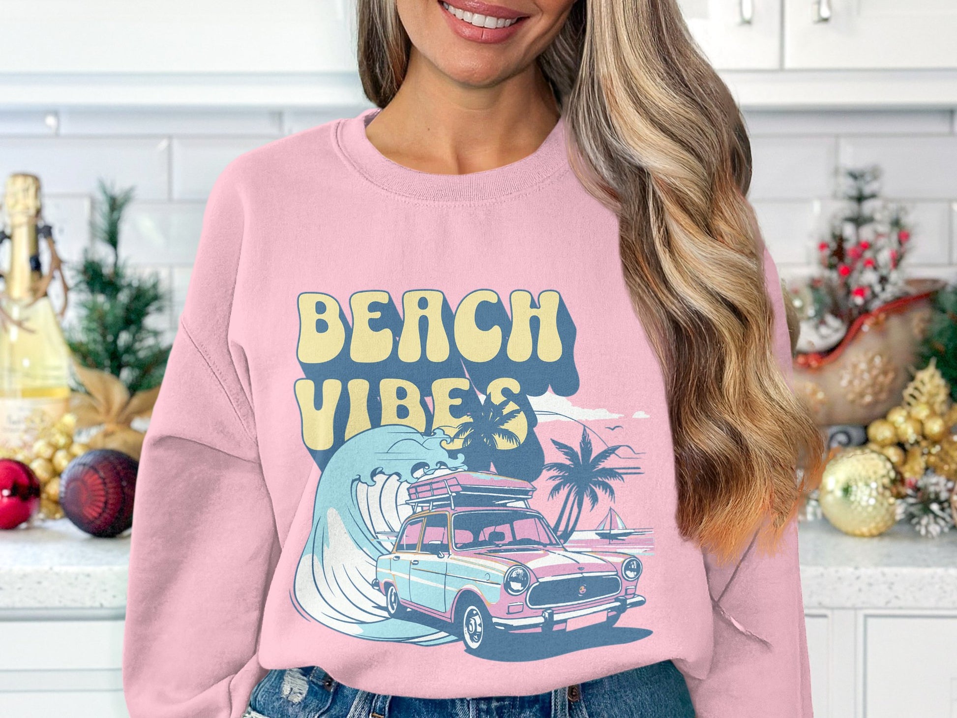 Wearing Garment Graphics pink retro sweatshirt, featuring a classic car, surfboard, wave, palm trees, and Beach Vibes, a person stands in a kitchen adorned with holiday ornaments and greenery.