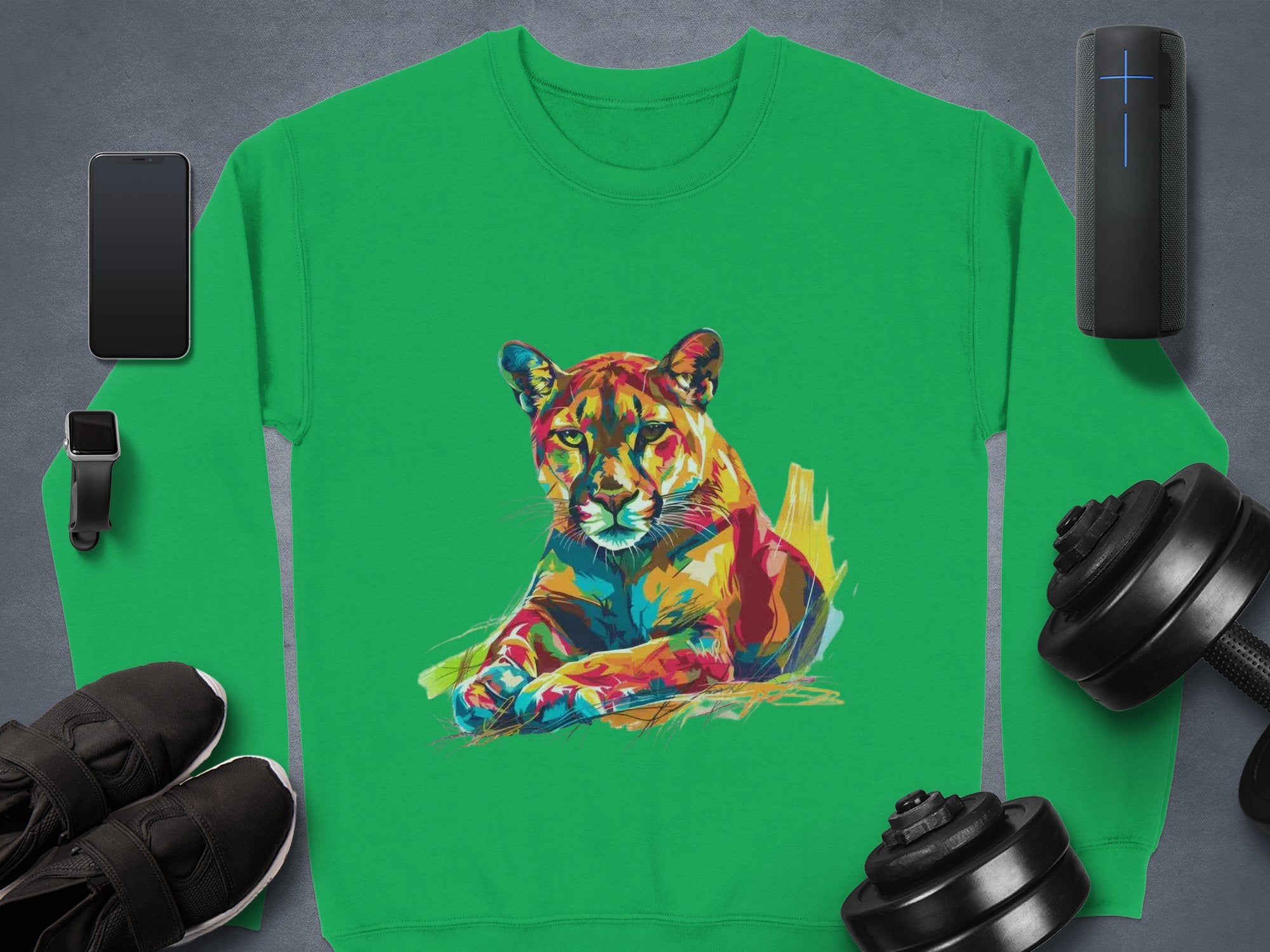 A Garment Graphics green sweatshirt with a colorful resting lioness design is displayed on a gray surface, surrounded by a smartphone, smartwatch, wireless speaker, shoes, and two adjustable dumbbells. Unisex sizing ensures it fits everyone.