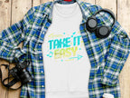 A Garment Graphics white t-shirt with the Take It Easy design in vibrant font is laid on a plaid shirt. Around it, a camera, headphones, and a coffee cup lid are artistically arranged on a wooden surface.
