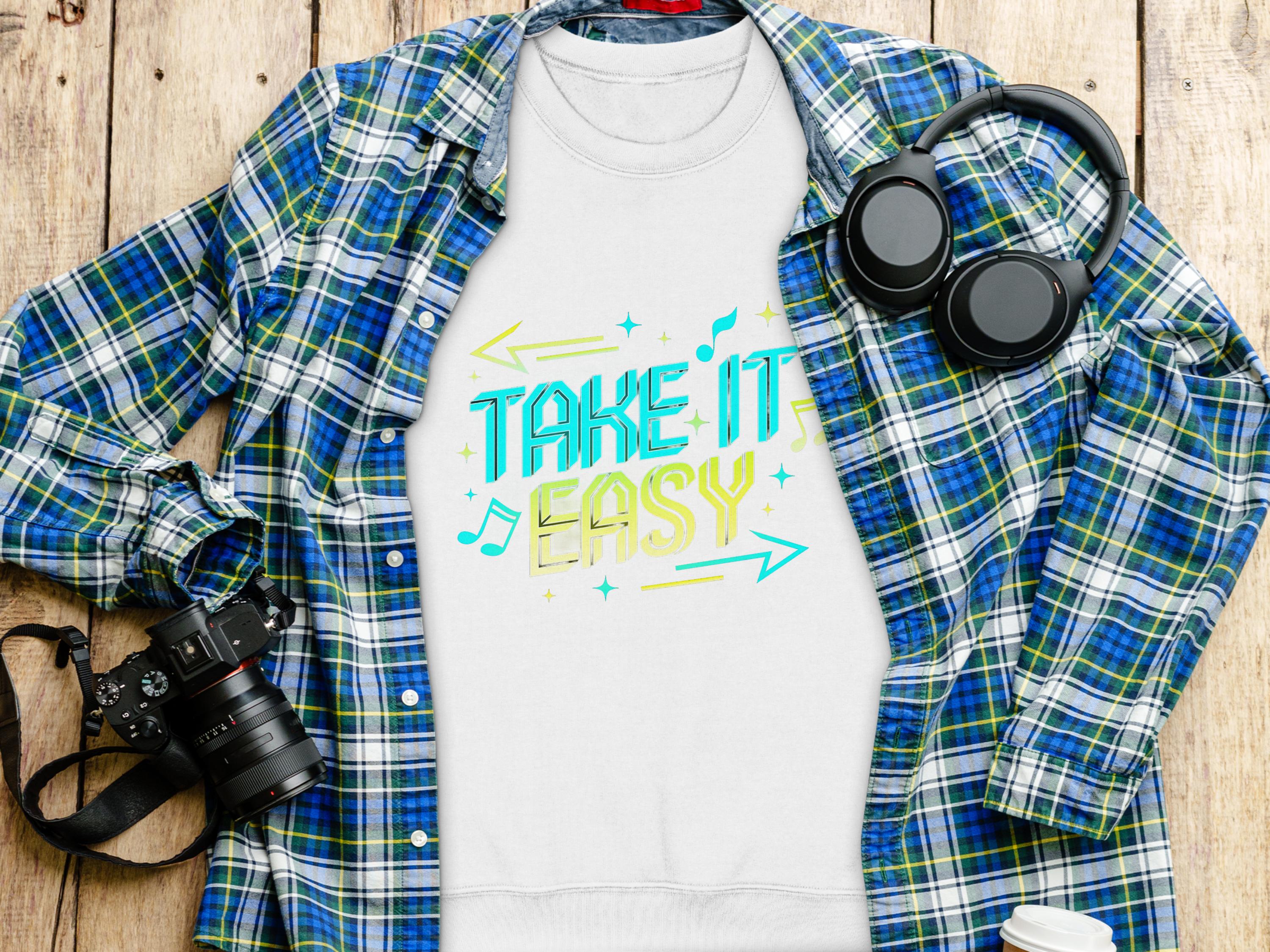 A Garment Graphics white t-shirt with the Take It Easy design in vibrant font is laid on a plaid shirt. Around it, a camera, headphones, and a coffee cup lid are artistically arranged on a wooden surface.