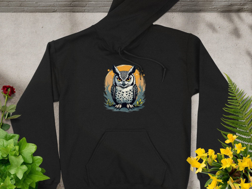 A Garment Graphics hoodie in black features a medium-heavy fabric with an owl wearing glasses, surrounded by leaves on a circular orange background, lying flat among green plants and yellow flowers, accentuating its classic fit elegance.