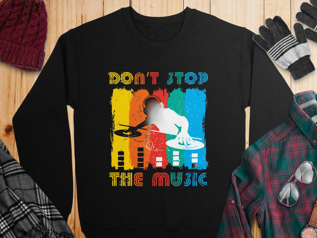 Garment Graphics presents a black sweatshirt for music lovers, featuring a vibrant DJ design with turntables and Dont Stop The Music boldly inscribed above and below. Displayed on a wooden surface, its surrounded by cozy winter clothing items.