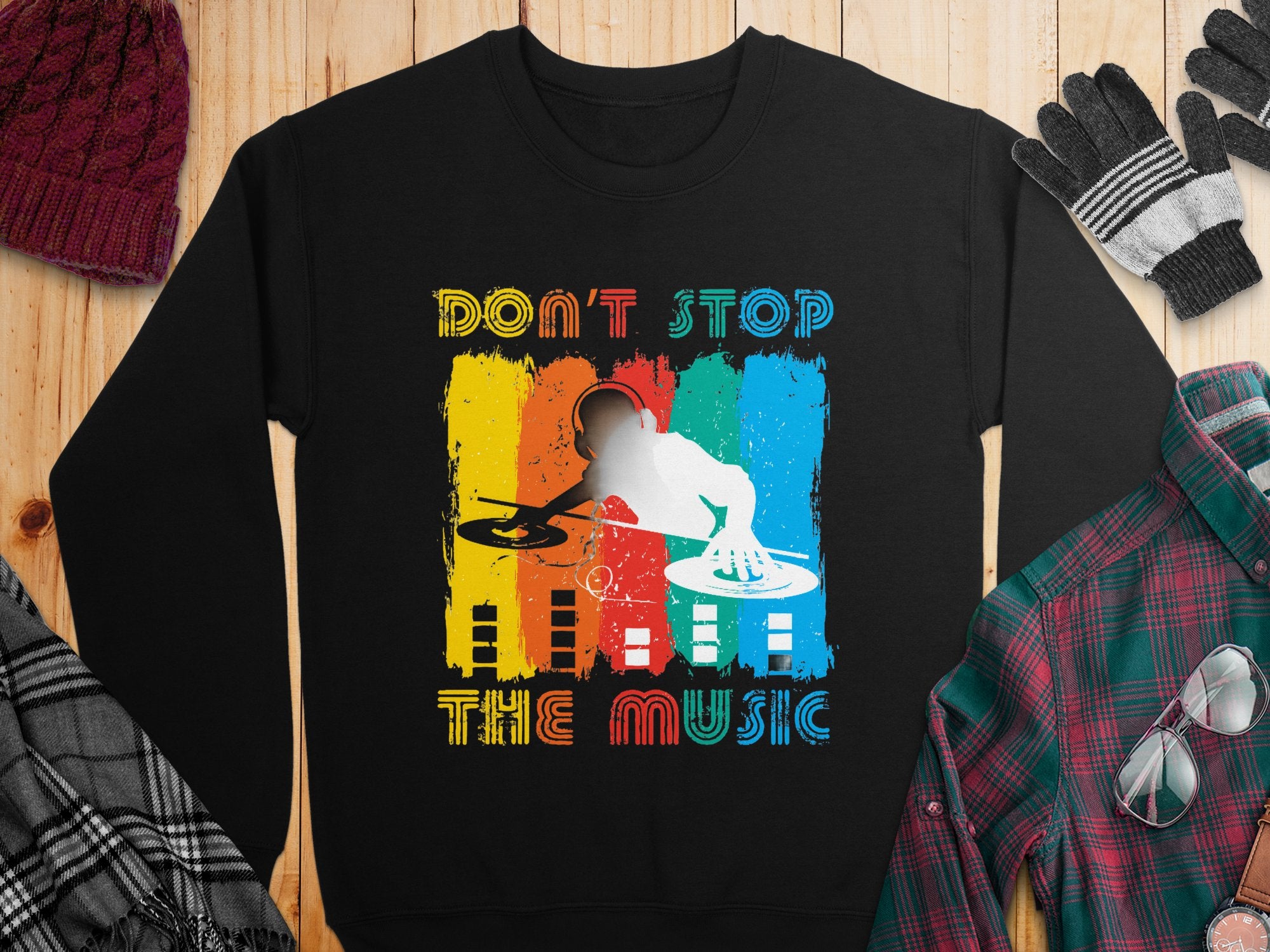 Garment Graphics presents a black sweatshirt for music lovers, featuring a vibrant DJ design with turntables and Dont Stop The Music boldly inscribed above and below. Displayed on a wooden surface, its surrounded by cozy winter clothing items.