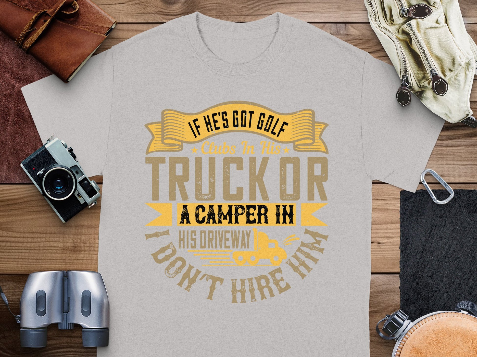 A witty gray t-shirt from Garment Graphics features a yellow and brown quote: If hes got golf clubs in his truck or a camper in his driveway, I dont hire him. Its the perfect gift, styled flat with a camera, binoculars, and leather notebook.