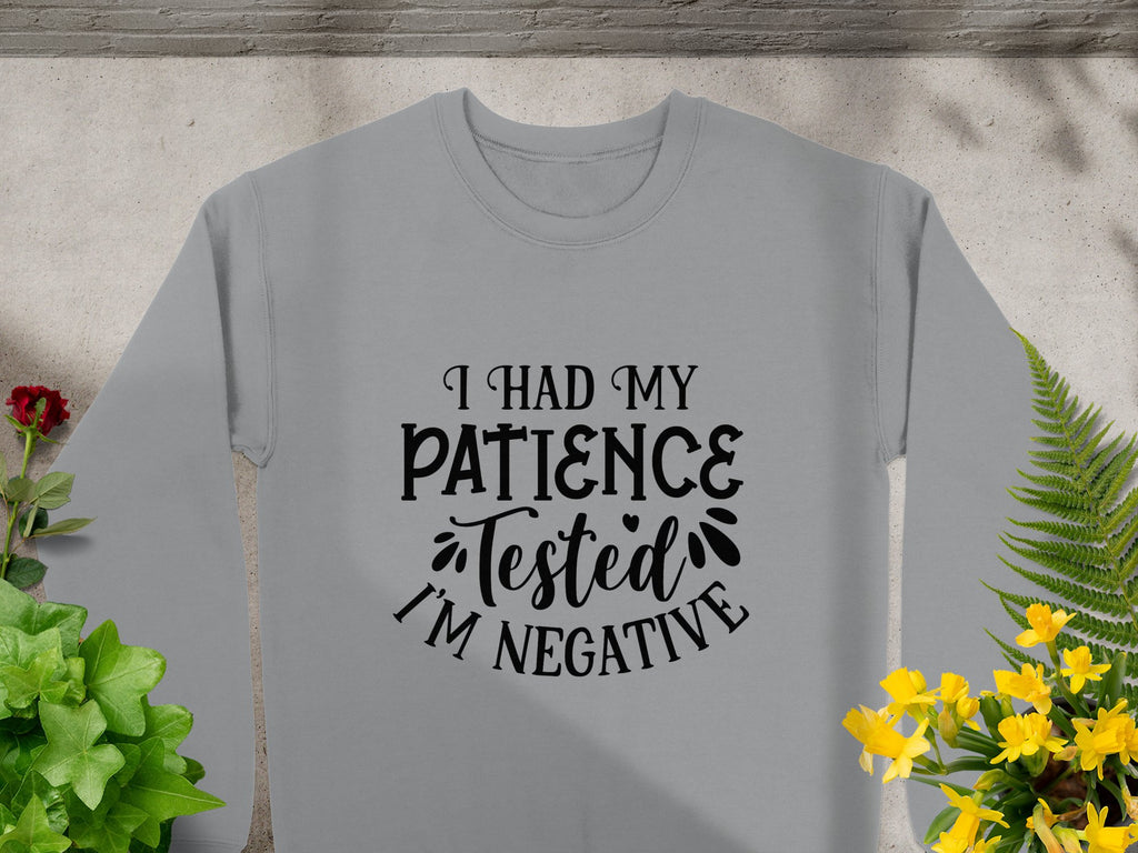 A Garment Graphics sweatshirt rests on concrete with green plants and yellow flowers. It features the phrase, I had my patience tested. Im negative, and decorative details. Ideal for casual outings, it offers unisex sizing for a comfortable fit for all.