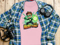 A pink T-shirt by Garment Graphics, showcasing a cartoon character and Its Hero Time!, adds playfulness to your wardrobe. Laid on a blue and green plaid shirt with a black camera, black headphones, and white cup lid on wood, its perfect for young heroes at heart!.