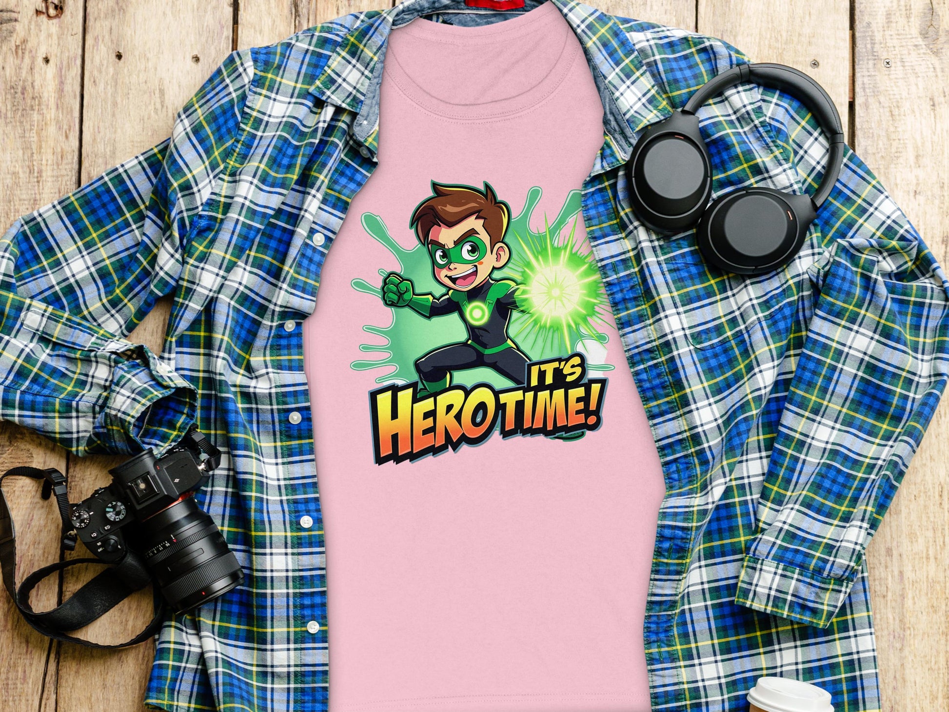 A pink T-shirt by Garment Graphics, showcasing a cartoon character and Its Hero Time!, adds playfulness to your wardrobe. Laid on a blue and green plaid shirt with a black camera, black headphones, and white cup lid on wood, its perfect for young heroes at heart!.