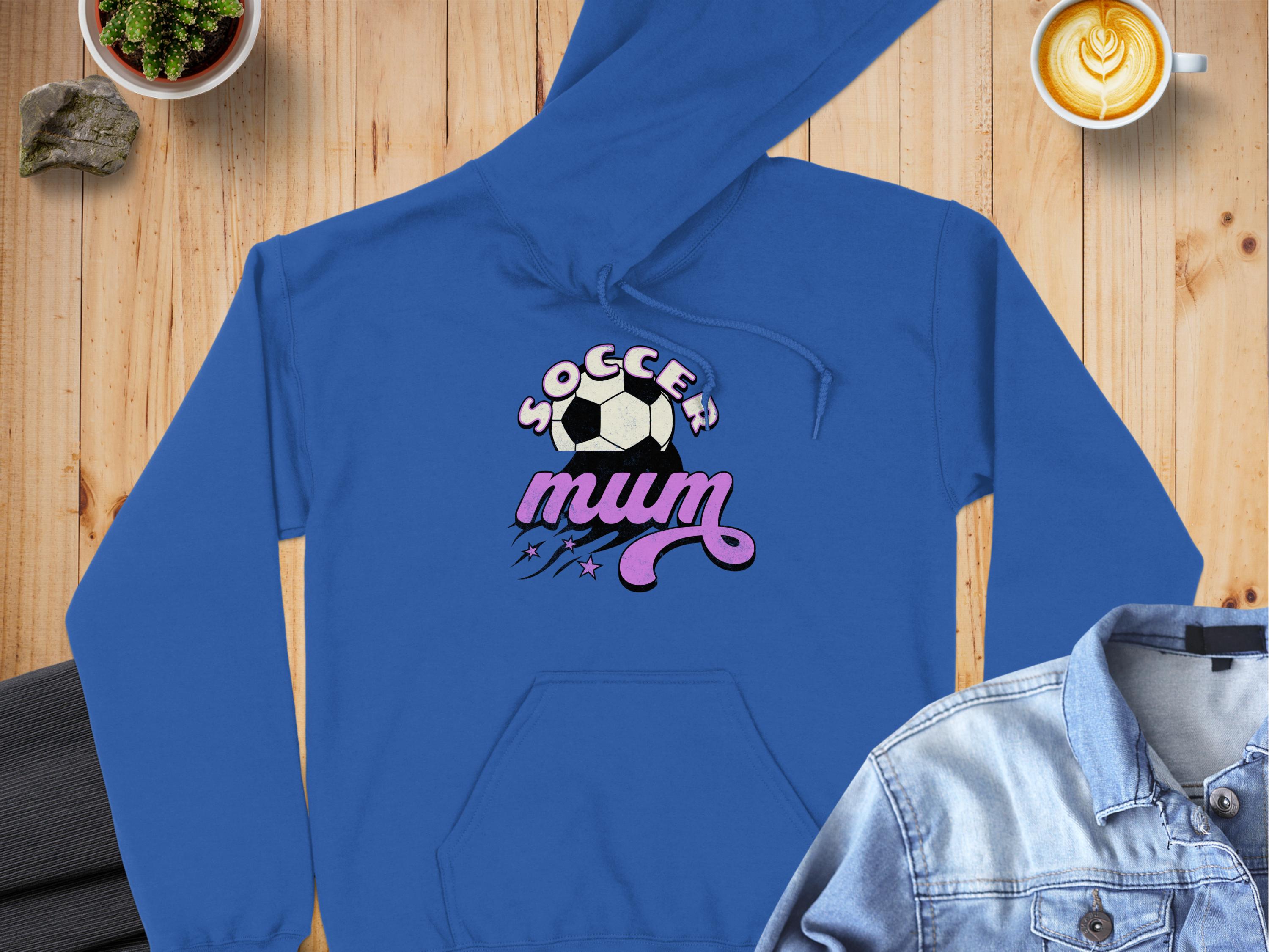 A cozy blue hoodie from Garment Graphics, featuring Soccer Mum and a soccer ball design in bold letters, lies on a wooden surface with a denim jacket, succulent plant, and artful latte.