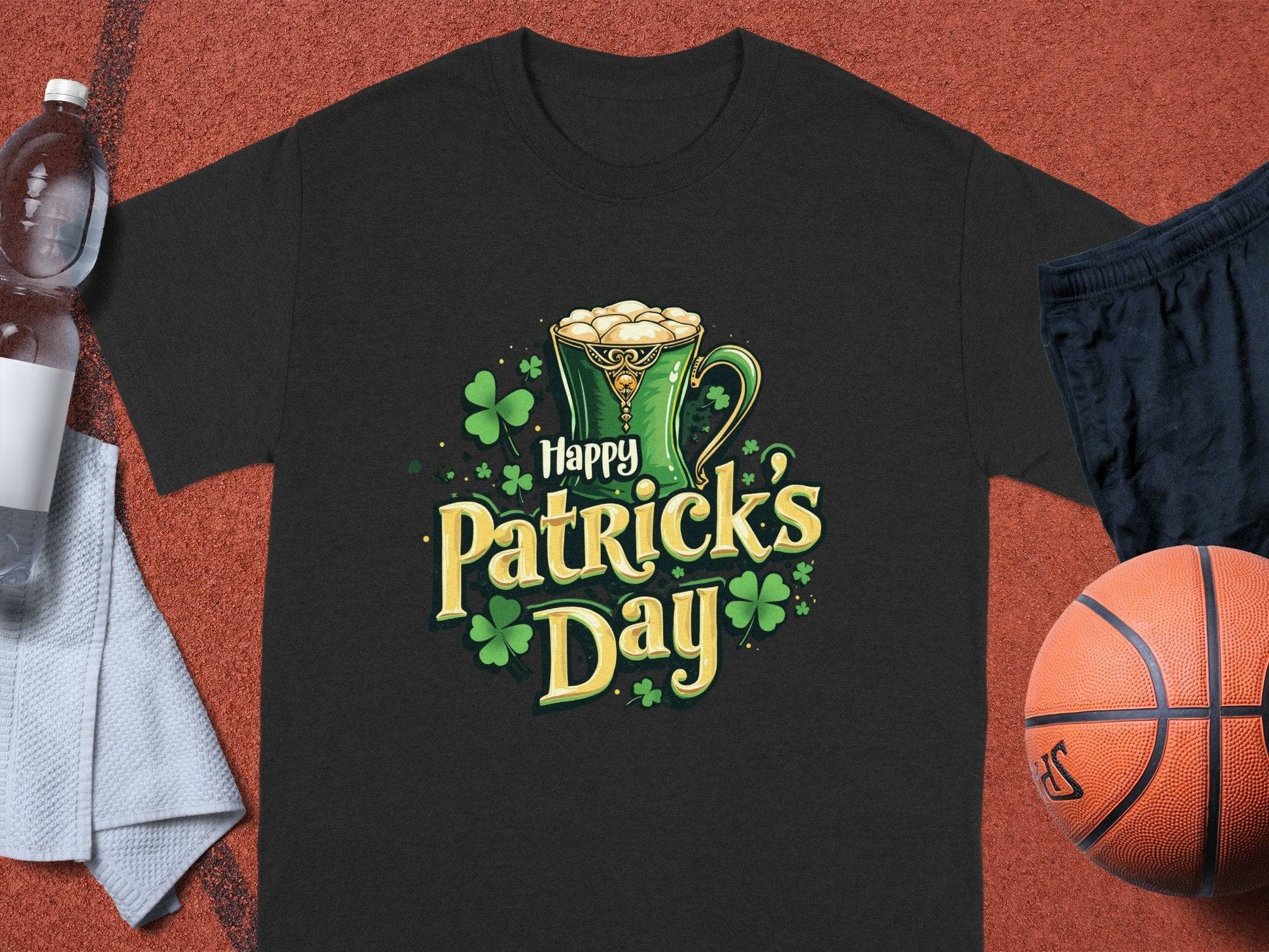 A festive black Happy Patricks Day T-shirt from Garment Graphics, featuring a green beer mug and shamrocks, sits on a red textured surface with a basketball, black shorts, white towel, and water bottle nearby.