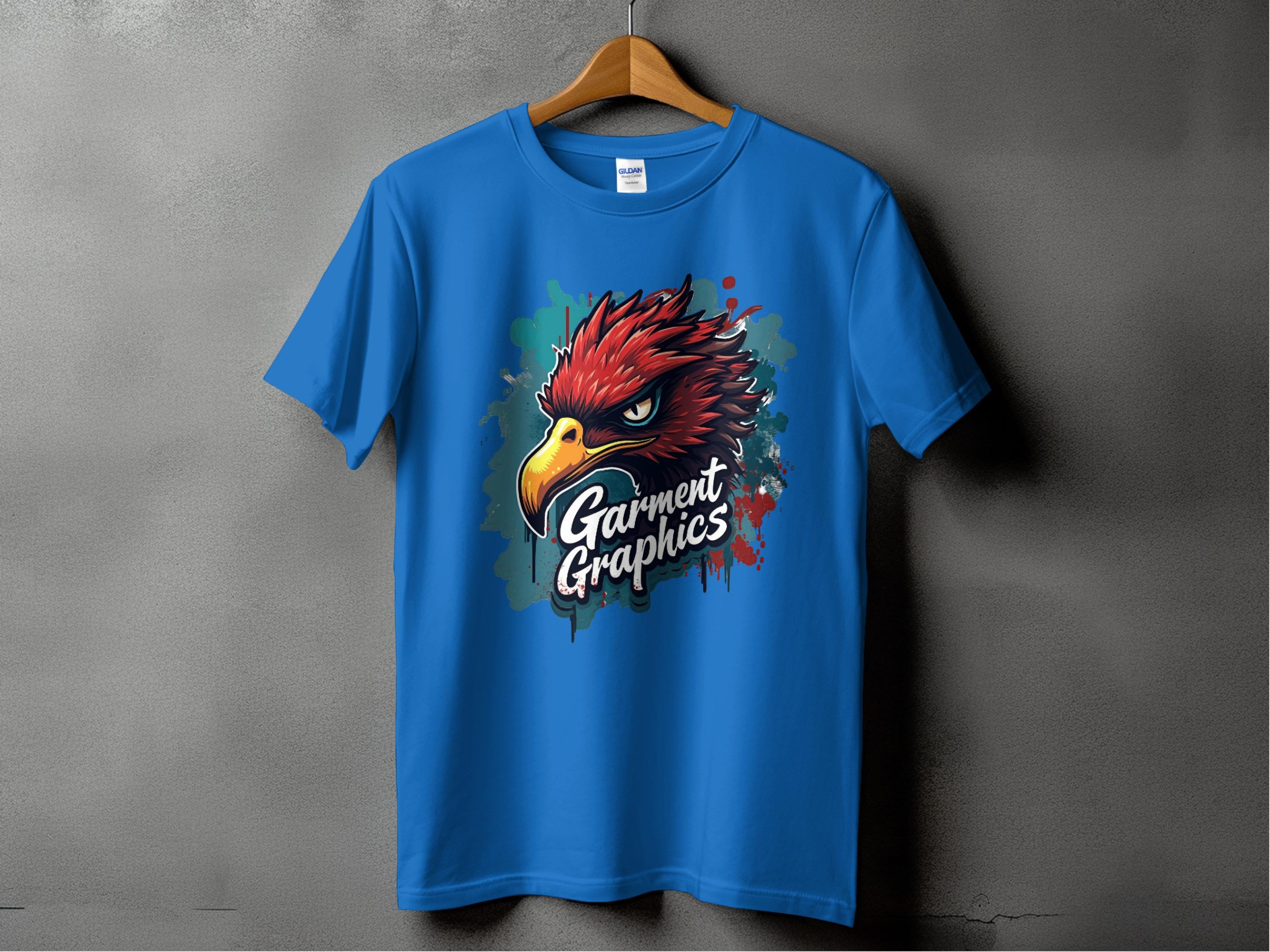 A blue T-shirt from Garment Graphics hangs on a gray wall, featuring a bold eagle design with fierce eyes. Garment Graphics is printed below the graphic, and a splash effect adds depth to this classic fit piece.