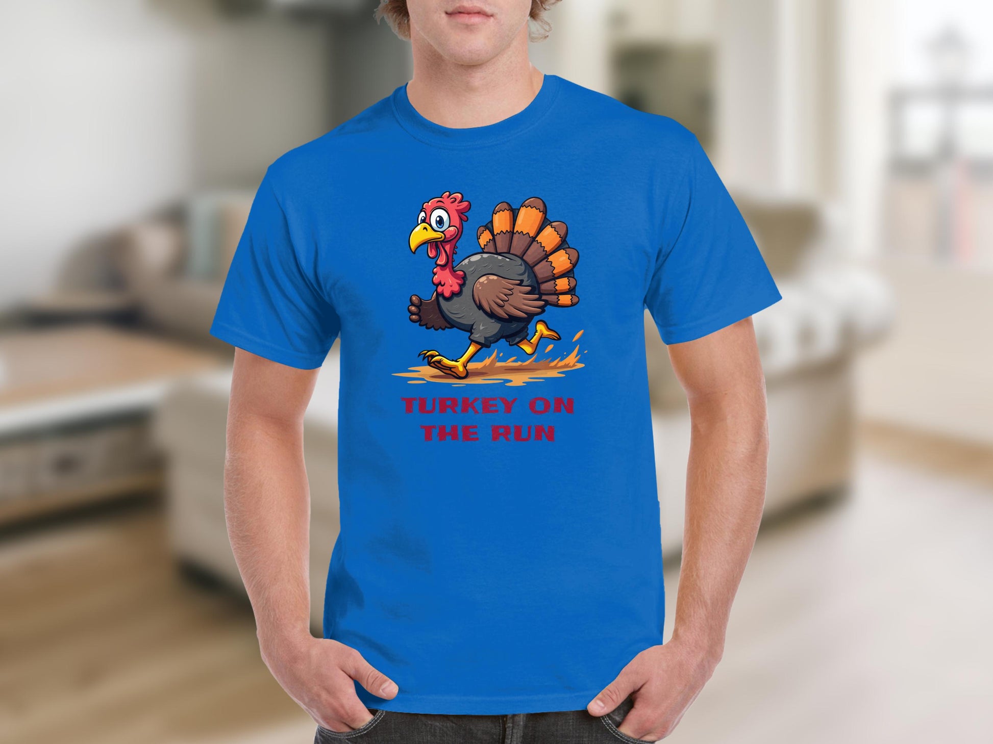 Someone is wearing a classic fit blue graphic T-shirt by Garment Graphics, featuring a cartoon turkey dashing across with the text TURKEY ON THE RUN. The scene is set in a blurred indoor background that resembles a cozy living room.