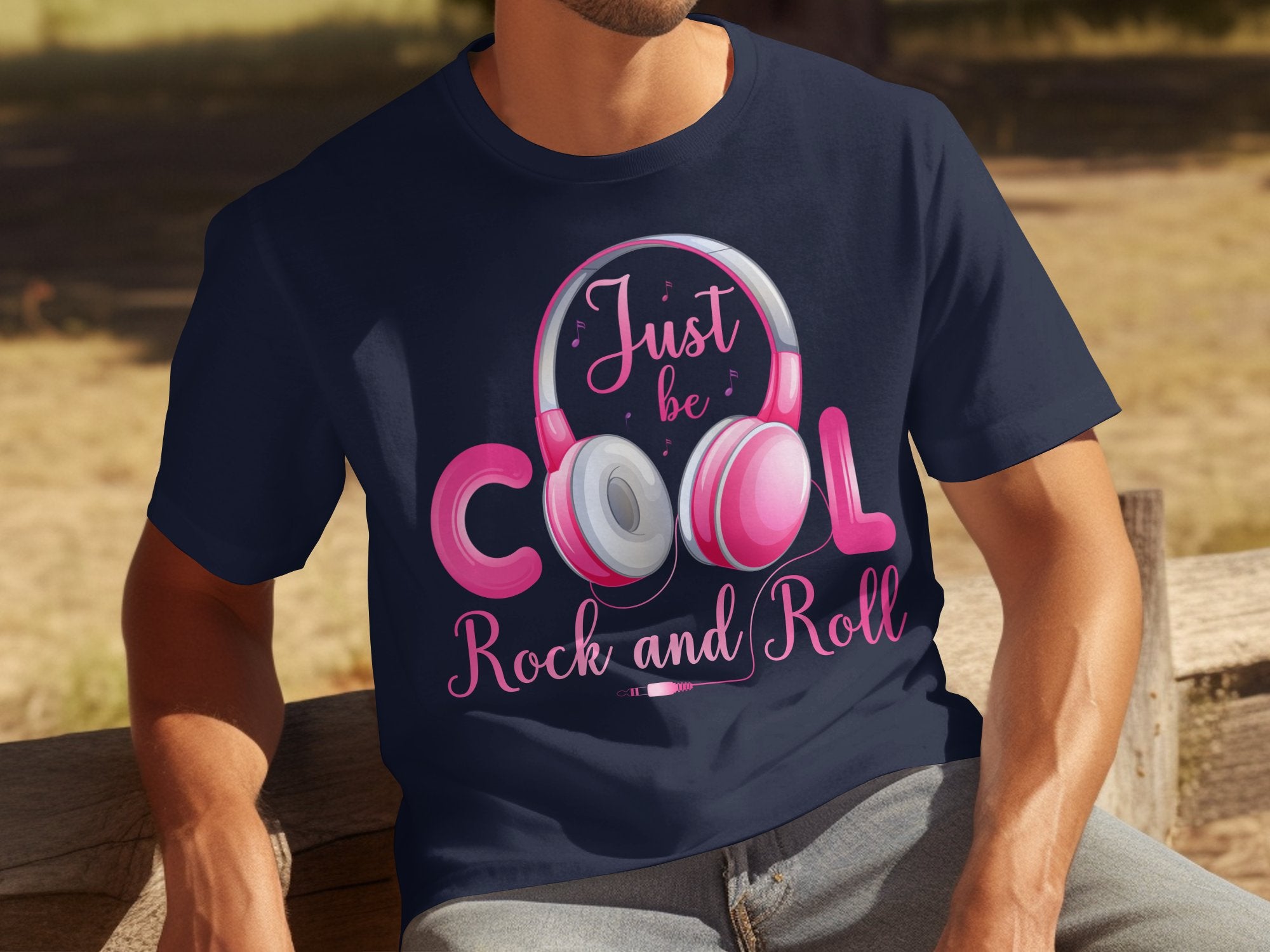 A person is sitting on a wooden bench outside, sporting a Garment Graphics navy blue T-shirt with pink and white headphones and the text Just be cool, Rock and Roll. Ideal for music enthusiasts wanting to make a statement.