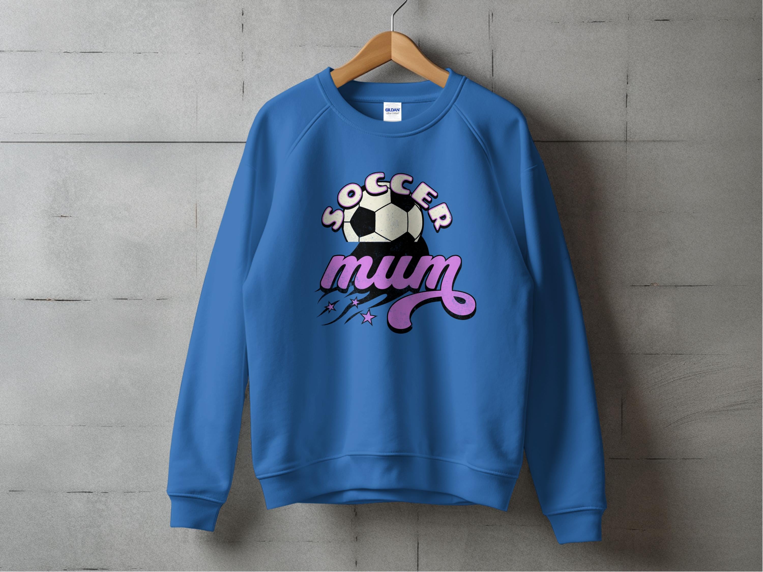 A vintage-style blue sweatshirt by Garment Graphics hangs on a wooden hanger against a gray concrete wall, featuring a soccer ball graphic with bold Soccer Mum text in white and pink, accented by stars.