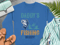 The Garment Graphics blue fishing sweatshirt features Daddys Fishing Buddy with a fish and boat image on a gray surface. It pairs well with a green purse, brown sandals, green striped shirt, white watch, and lush green ferns—ideal for fishing enthusiasts.