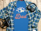 A Soccer Dad blue graphic T-shirt from Garment Graphics, featuring a soccer ball design, is placed on a plaid shirt on wood. Nearby are headphones, a camera, a notebook, and a coffee cup lid.