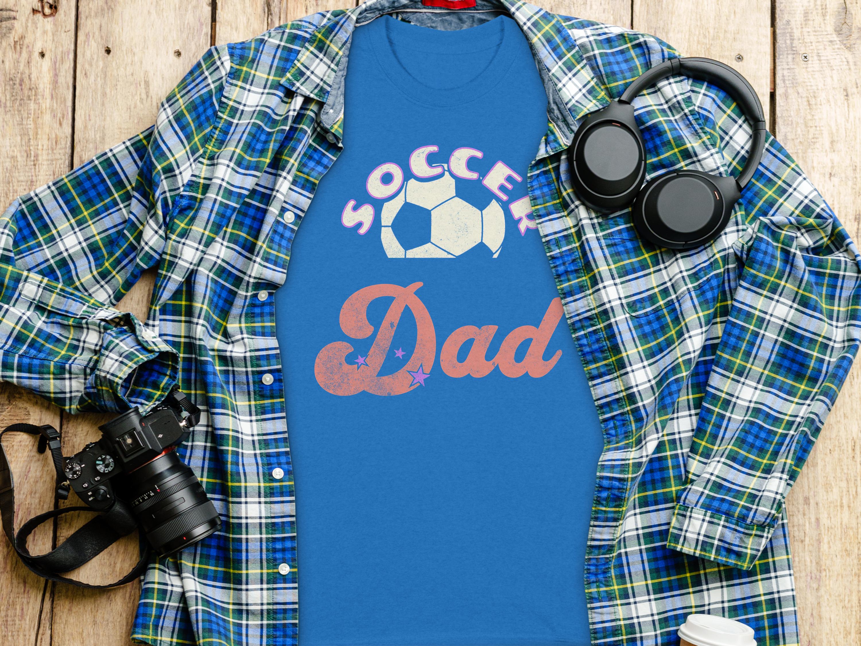 A Soccer Dad blue graphic T-shirt from Garment Graphics, featuring a soccer ball design, is placed on a plaid shirt on wood. Nearby are headphones, a camera, a notebook, and a coffee cup lid.