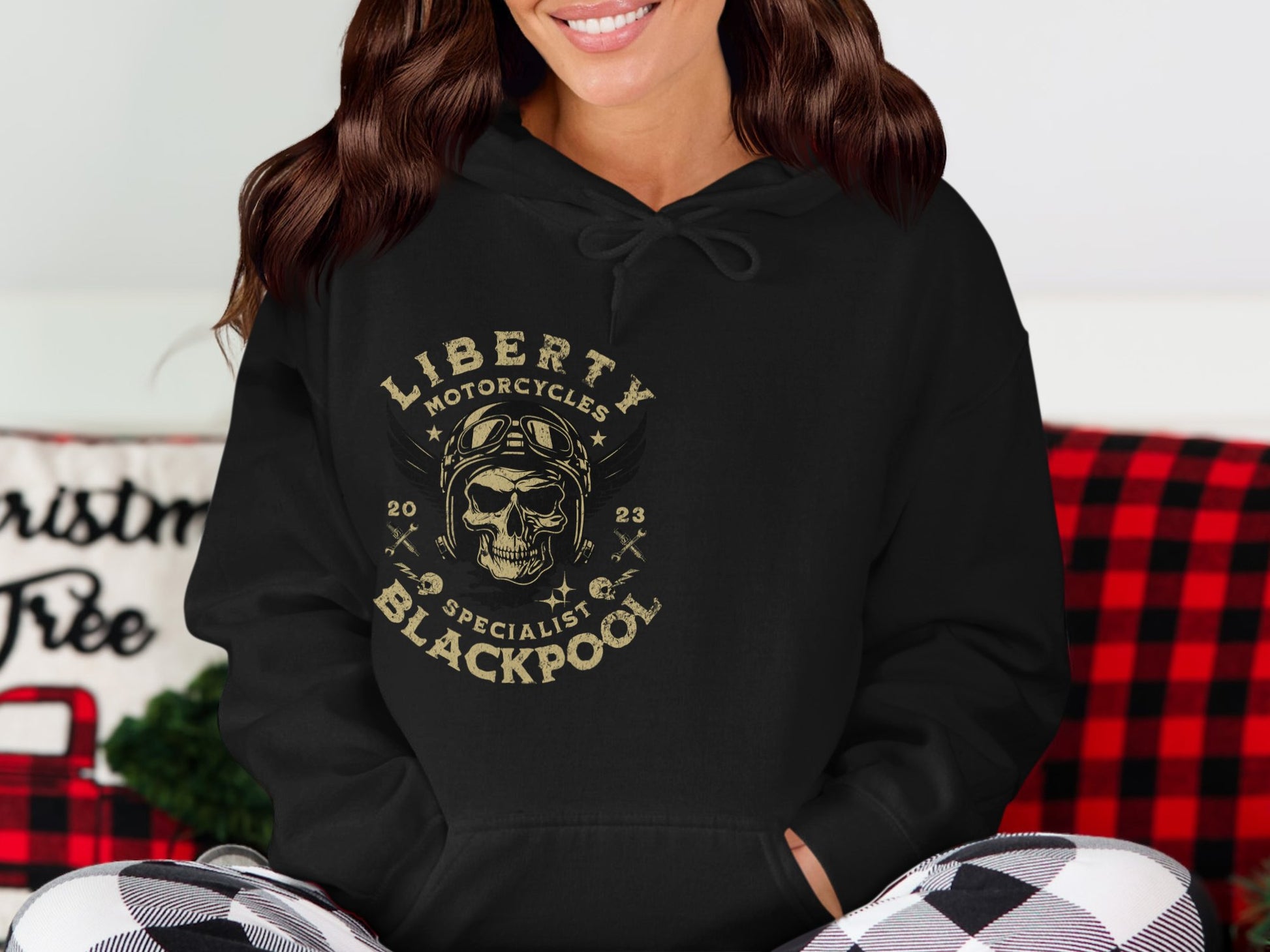 Wearing the Garment Graphics Blackpool Hoodie, featuring a black design with a skull, goggles, wings, and Liberty Motorcycles Specialist Blackpool 2023 text. In the background is a checkered red blanket and part of a Christmas tree sign.