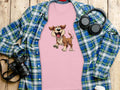 A dog lovers pink T-shirt from Garment Graphics, featuring a cartoon dog print, is spread over a blue and green plaid shirt. Nearby are a camera, headphones, and a take-out cup with lid, all placed on a wooden surface.