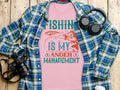 A Garment Graphics pink fishing T-shirt, featuring Fishing Is My Anger Management, rests on wood beside a plaid shirt, headphones, camera, and white reusable bottle—ideal for the relaxed style of an avid fishing enthusiast.