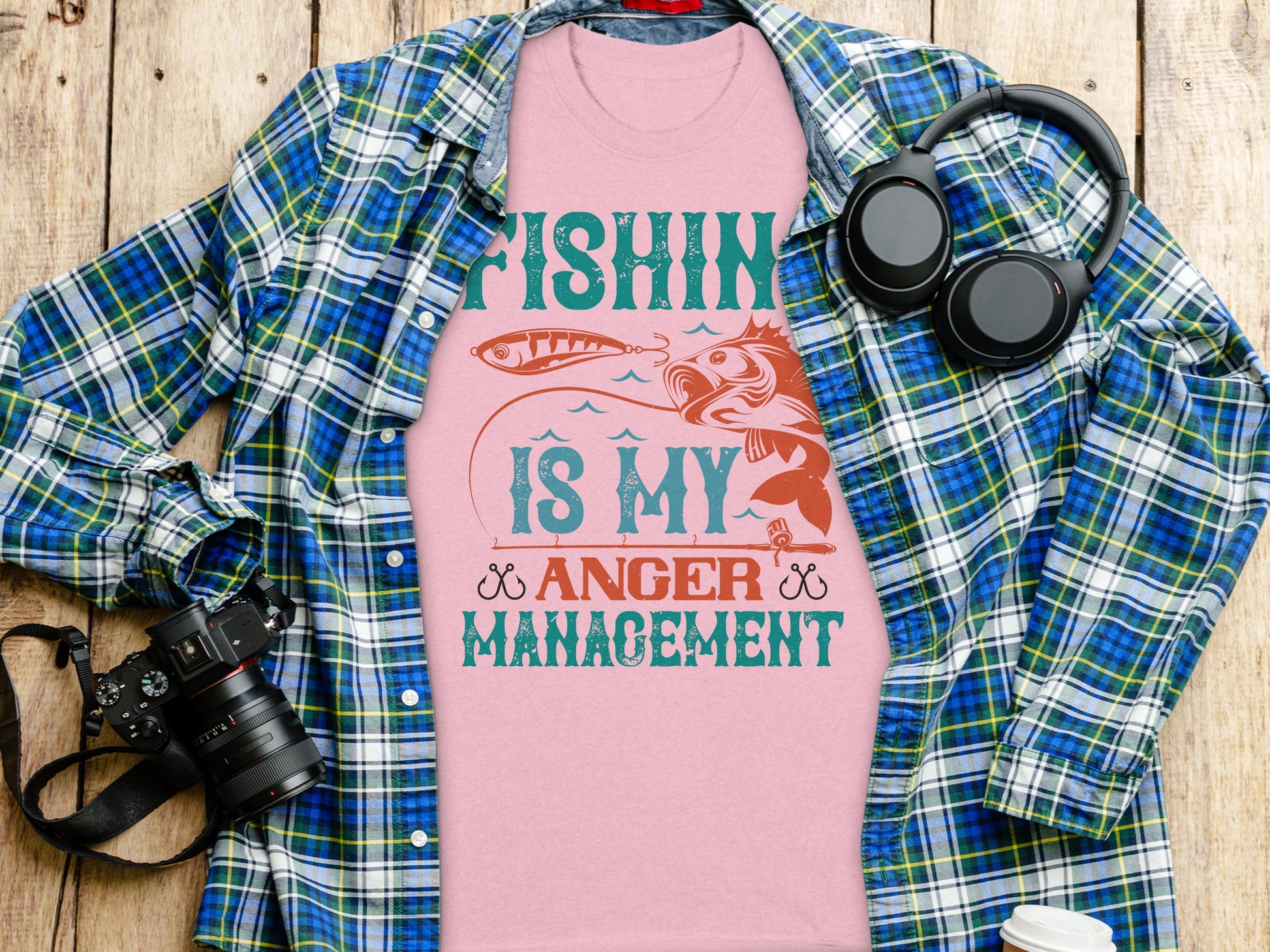 A Garment Graphics pink fishing T-shirt, featuring Fishing Is My Anger Management, rests on wood beside a plaid shirt, headphones, camera, and white reusable bottle—ideal for the relaxed style of an avid fishing enthusiast.