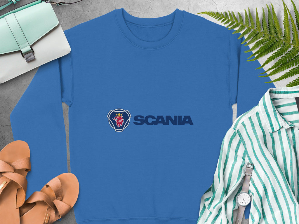 A blue unisex Scania logo sweatshirt by Garment Graphics is displayed on a surface, surrounded by sandals, a striped shirt, a wristwatch, a green purse, and green fern leaves—ideal for casual outings.