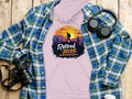 A pink Garment Graphics hoodie showcases a hiking silhouette with Retired 2025, Not My Problem Anymore over a sunset mountain design on a wooden surface, accompanied by a plaid shirt, headphones, and camera.