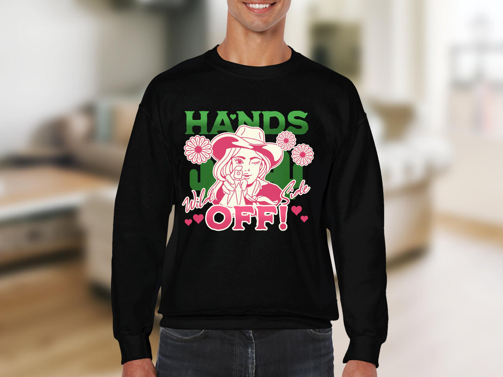A man wears a black Garment Graphics Wild Side Sweatshirt featuring bold cartoon character graphics, including a flowered hat and the text Hands off! and Wild girl. This unisex sweatshirt stands out against the blurred indoor backdrop.