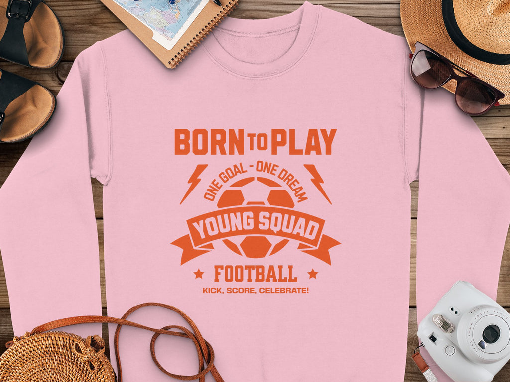 The Garment Graphics pink sweatshirt features an orange soccer ball design with the text: BORN TO PLAY. ONE GOAL - ONE DREAM. YOUNG SQUAD FOOTBALL. Ideal for young squads, this unisex piece is surrounded by a hat, sunglasses, notebook, and shoes on a wooden surface.