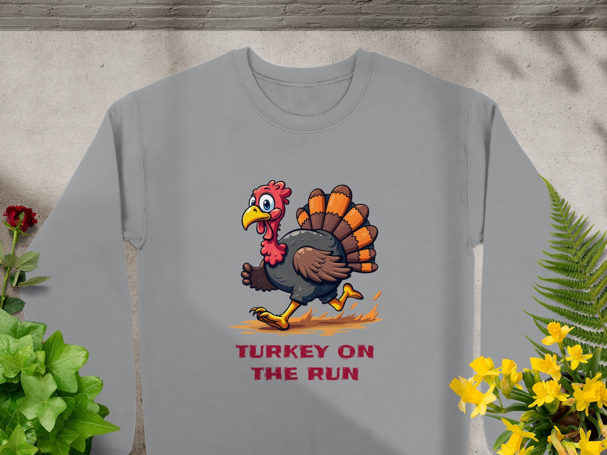 The Garment Graphics gray sweatshirt, designed for unisex sizing, showcases a humorous cartoon turkey on the run. Its complemented by green and yellow plants and a red flower on the left, adding charm to this funny graphic design.