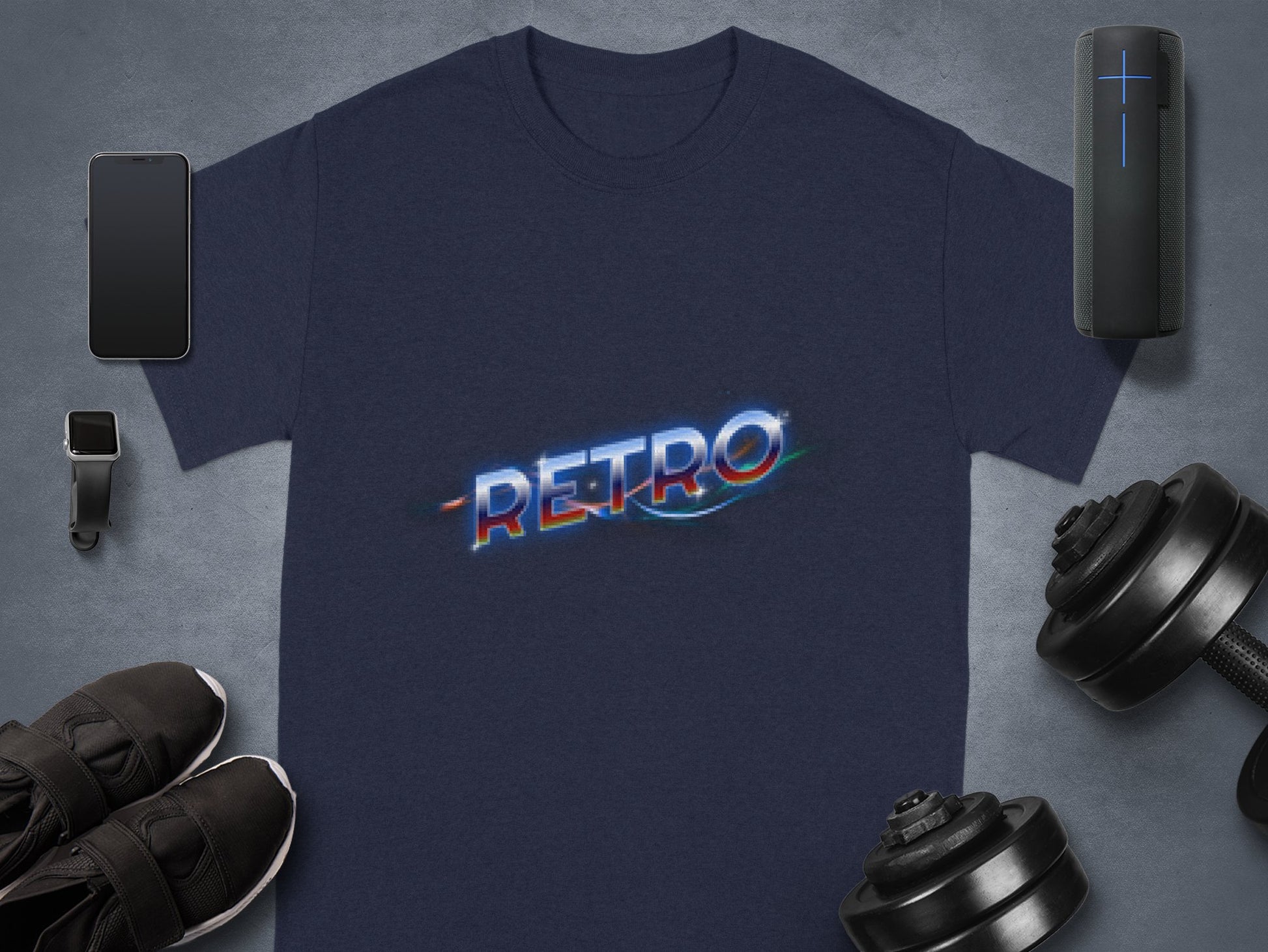 A navy blue T-shirt with a multicolored RETRO graphic from Garment Graphics exudes vintage charm. The classic fit piece is surrounded by a smartphone, smartwatch, black sneakers, portable speaker, and adjustable dumbbells on a gray surface.