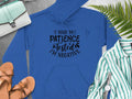 The Garment Graphics Blue Patience Tested Hoodie features black text saying I had my patience tested, Im negative with fern leaves. It’s a classic fit displayed with a green handbag, striped shirt, brown sandals, and watch on a gray background.