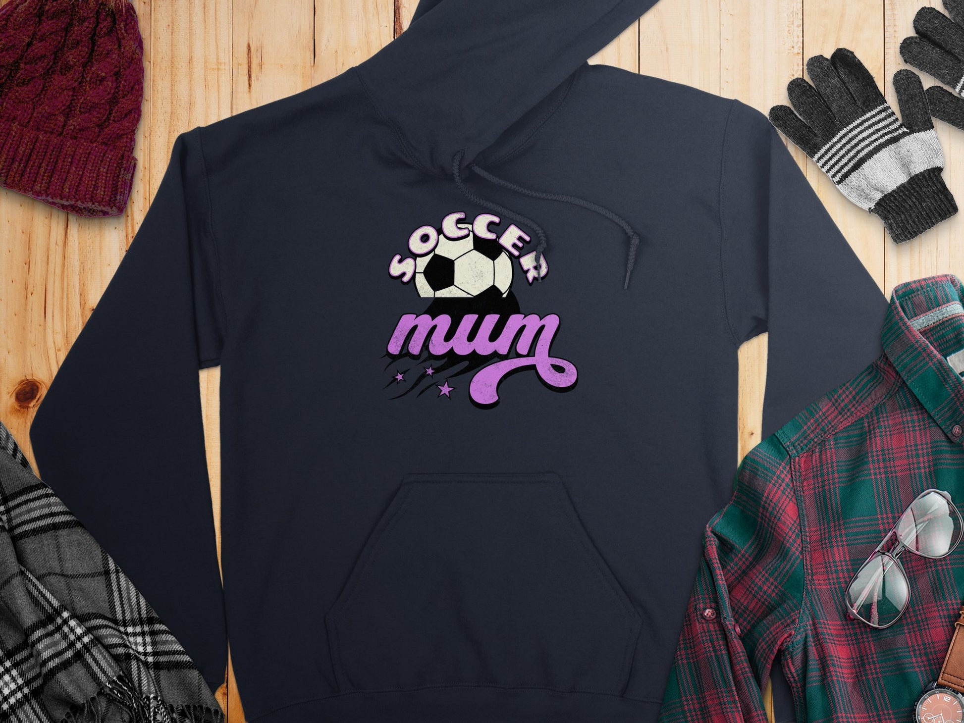 A navy Soccer Mum Hoodie from Garment Graphics, featuring a classic fit and soccer ball graphic, rests on wood accompanied by a maroon knit hat, gray and black striped gloves, a plaid shirt, and a fringed scarf.