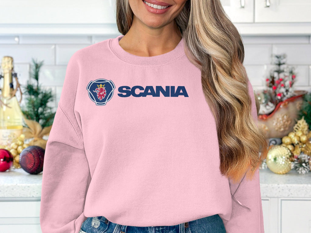A woman with wavy blonde hair smiles in a kitchen wearing a light pink Garment Graphics Scania sweatshirt, ideal for casual outings. The unisex sizing offers comfort for all. Behind her, the counter is decorated with Christmas ornaments and small trees.