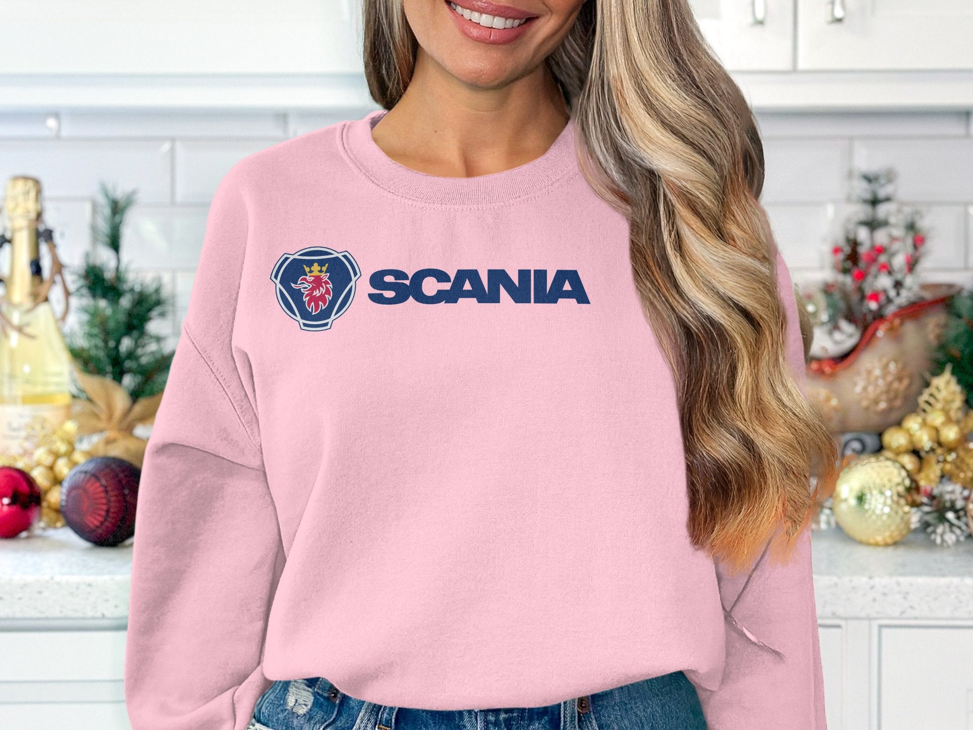 A woman with wavy blonde hair smiles in a kitchen wearing a light pink Garment Graphics Scania sweatshirt, ideal for casual outings. The unisex sizing offers comfort for all. Behind her, the counter is decorated with Christmas ornaments and small trees.