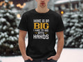 In a snowy setting, someone stands with hands in pockets wearing a Garment Graphics classic fit tee. The black shirt humorously reads, “Mine is so big I have to use both hands,” with certain words highlighted in playful colors.