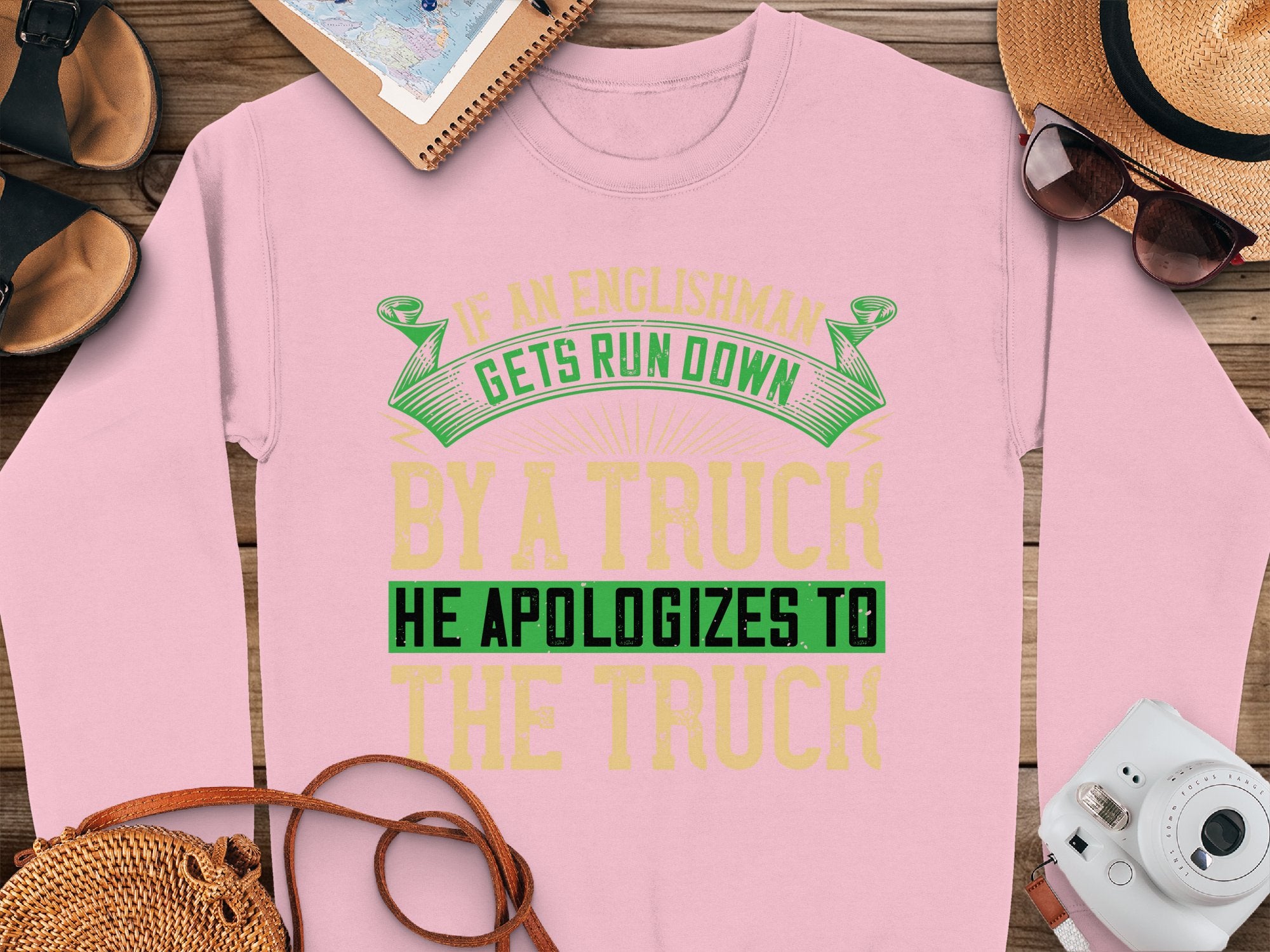 This pink Garment Graphics sweatshirt features a bold, humorous design with green and yellow text: If an Englishman gets run down by a truck he apologizes to the truck. Its unisex-sized and complements well with accessories like a hat, sunglasses, journal, watch, or woven bag.
