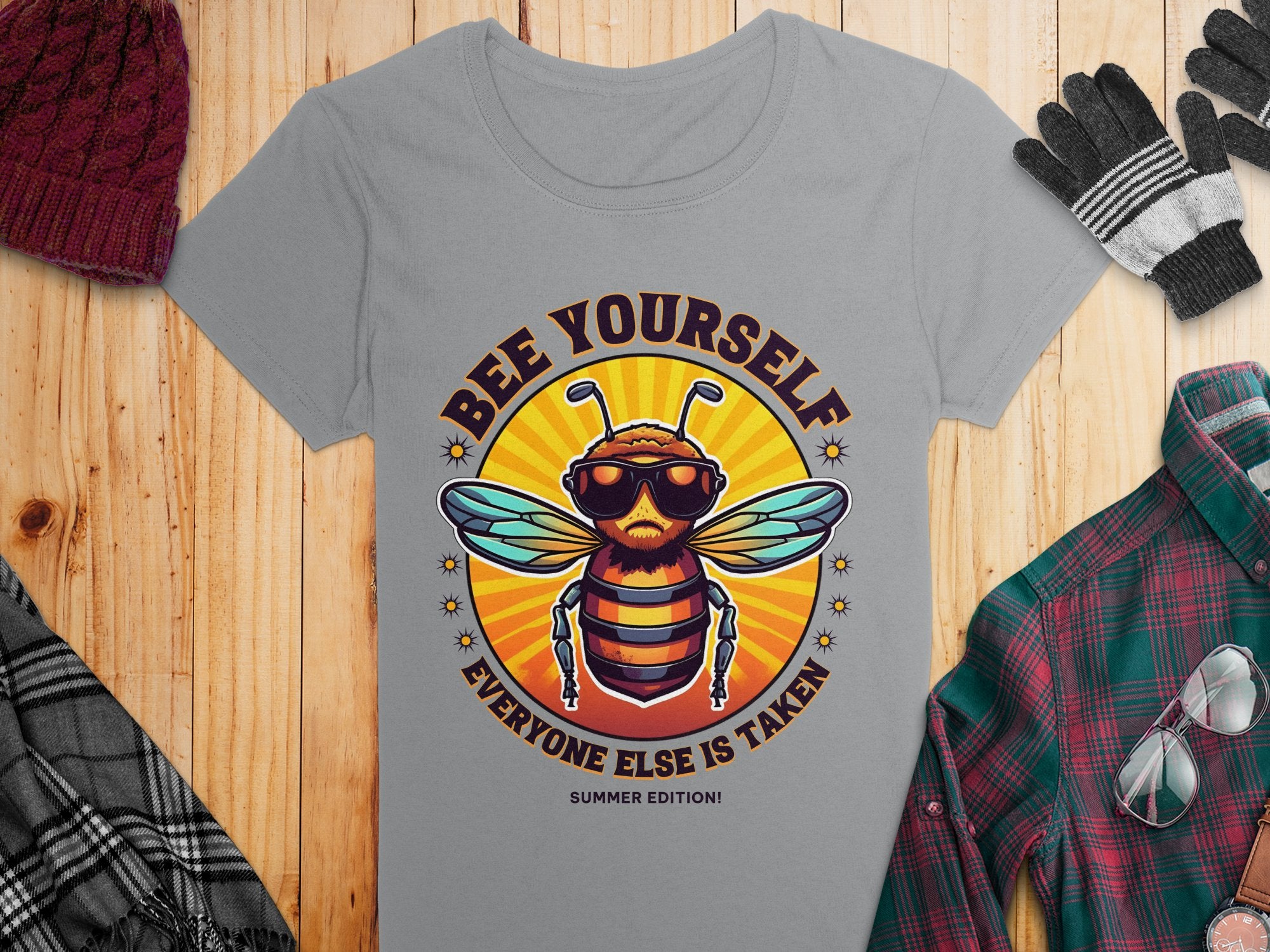Discover Garment Graphics playful Bee Yourself T-Shirts, Summer Edition! This gray womens tee showcases a bee in sunglasses and headphones, accompanied by a red knit cap, black gloves, plaid shirt, and sunglasses on a wooden surface.