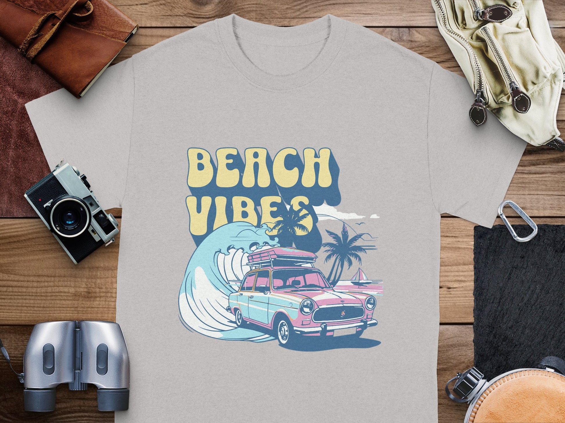 Garment Graphics presents a gray graphic t-shirt featuring a classic car with a surfboard, a large wave, and palm trees. BEACH VIBES is printed above the image. Its artistically displayed on wood, surrounded by travel accessories.