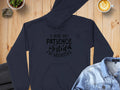 A black Garment Graphics Patience Tested Hoodie with the text, I had my patience tested, Im negative, sits on a wooden table next to a latte, a small plant, folded pants, and a denim shirt. This classic fit hoodie is made from medium-heavy fabric for comfort.